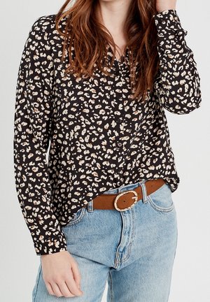 Femme portant un chemisier noir à manches longues avec un motif floral abstrait blanc et marron, rentré dans un jean bleu clair avec une ceinture marron.