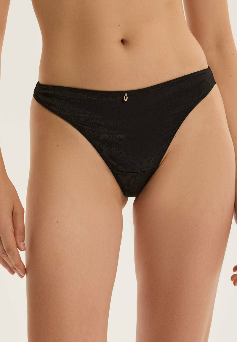Schwarzer Spitzen-Thong mit einem strukturierten Muster und einem goldfarbenen Akzent am Bund. Weicher Stoff mit einem tief geschnittenen Schnitt.