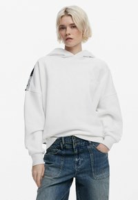 Hvid hoodie med en front kanguru lomme, ribbede manchetter og et subtilt præget design på brystet, båret med slidte denim jeans.