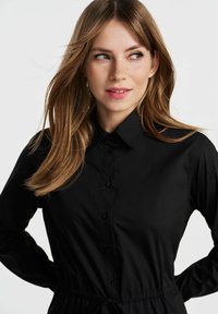 Czarna koszula z guzikami, z kołnierzykiem, wysmuklona w pasie i z długimi rękawami. Gładka tekstura i elastyczny sznurek do regulacji dopasowania.