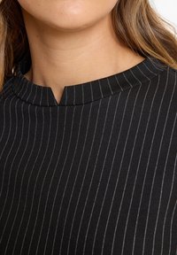 Zwarte shirt met verticale witte strepen, met een hoge, ronde halslijn en een kleine inkeping aan de voorkant in het midden. Gladde stoftextuur.