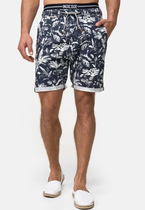Shorts de baño de hombre en azul marino con un patrón de hojas tropicales en blanco, fabricados con tejido, con cintura ajustable y dobladillo enrollado. Combinados con zapatillas blancas.