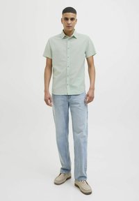 Camisa corta de manga ligera verde claro con botón frontal, combinada con jeans azul claro y zapatillas beis sin cordones. Tejido suave, corte regular.