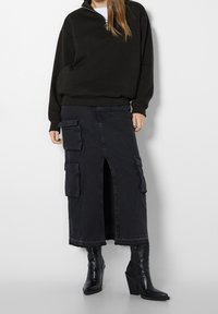 Femme portant un pull noir à manches longues, une jupe midi cargo noire avec une fente frontale, et des bottes noires à talons à bout pointu sur un fond uni.