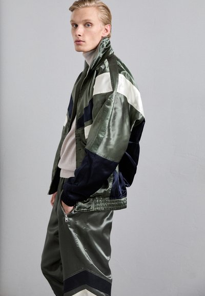 Veste bomber en satin vert avec des accents bleu marine et crème, accompagnée d'un pantalon assorti arborant un motif géométrique. Texture lisse et brillante.