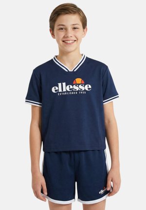 Enfant souriant portant un t-shirt ellesse bleu marine et un short assorti avec une bordure blanche, debout devant un fond clair uni.