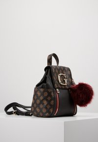 Sac à dos en similicuir noir avec accents marron à motif, ferrures argentées, rabat avant et détail pompon en fausse fourrure bordeaux.