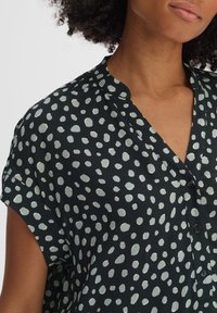 Femme portant un blouse noire à manches courtes avec des pois irréguliers blancs et un col mandarin, boutons partiellement fermés.