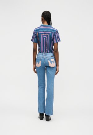 Személy fonott hajjal, lila és kék csíkos rövid ujjú ingben, világoskék farmernadrágban, melynek hátsó zsebein rózsaszín lángminták találhatók.