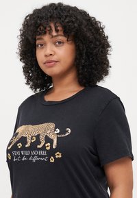Svart bomull t-skjorte med et leopardmotiv med gullfarge. Designet inkluderer tekst som står "Vær vill og fri, men vær annerledes."