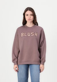 Preveliki ljubičasti dukserica s natpisom "BLUSH" u podebljanim bež slovima. Ima okrugli izrez i rebraste manžete. U kombinaciji s plavim farmerkama.