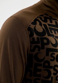 Braunfarbenes Langarmoberteil mit einem großen schwarzen Textmuster auf einer Seite, aus glattem Stoff mit kontrastierenden Nähdetails.