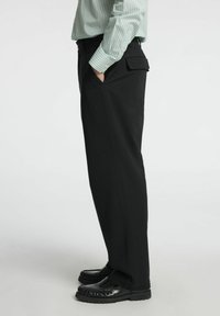 Juodos pritaikytos kelnės, pagamintos iš tekstūruoto audinio, su šoniniais kišenėmis ir tiesiu kirpimu, derinamos su juodais mokasinais.