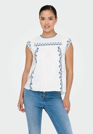 Witte blouse met blauwe borduursels langs de halslijn en zijkanten, met korte mouwen en een ontspannen pasvorm. Gecombineerd met blauwe spijkerbroeken.