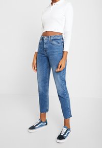 Avkortad vit långärmad polo skjorta i kombination med högmidjade, blekta blå jeans med fransade kanter, bärs med marinblå sneakers.