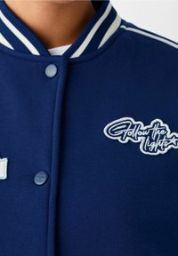 Mörkblå varsityjacka med vit randig krage, tryckknappar och broderad "Follow the Lights"-patch. Mjuk tygstruktur.