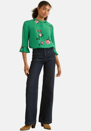 Groene bloemenblouse met puffmouwen, gecombineerd met hoge donkerdenim flared broeken en tan haklaarzen. Textuur en accenten zichtbaar.