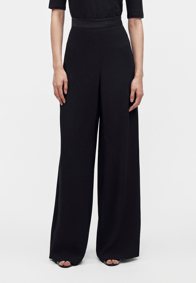 Personne portant un pantalon noir taille haute et jambes larges, un haut noir à manches au coude et des chaussures noires ouvertes, debout sur fond blanc.