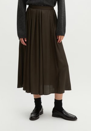 Femme portant une jupe midi plissée olive foncé, des chaussettes noires, des mocassins en cuir noir et un pull à manches longues gris foncé, debout sur fond blanc.
