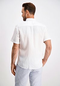 Camisa blanca de lino de manga corta con un cuello clásico, que presenta un pliegue en la parte posterior y puños enrollados, combinada con pantalones claros a rayas.