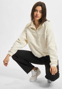 Cremfarbener Zip-Up-Sweatshirt mit strukturellem Kontrast über der Brust, kombiniert mit schwarzen, cropped Jeans und weißen Sneakers mit goldenen Akzenten.