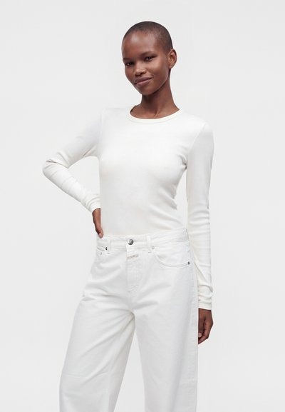 Personne aux cheveux courts portant une chemise blanche ajustée à manches longues et un jean blanc, debout avec une main sur la hanche devant un fond uni.