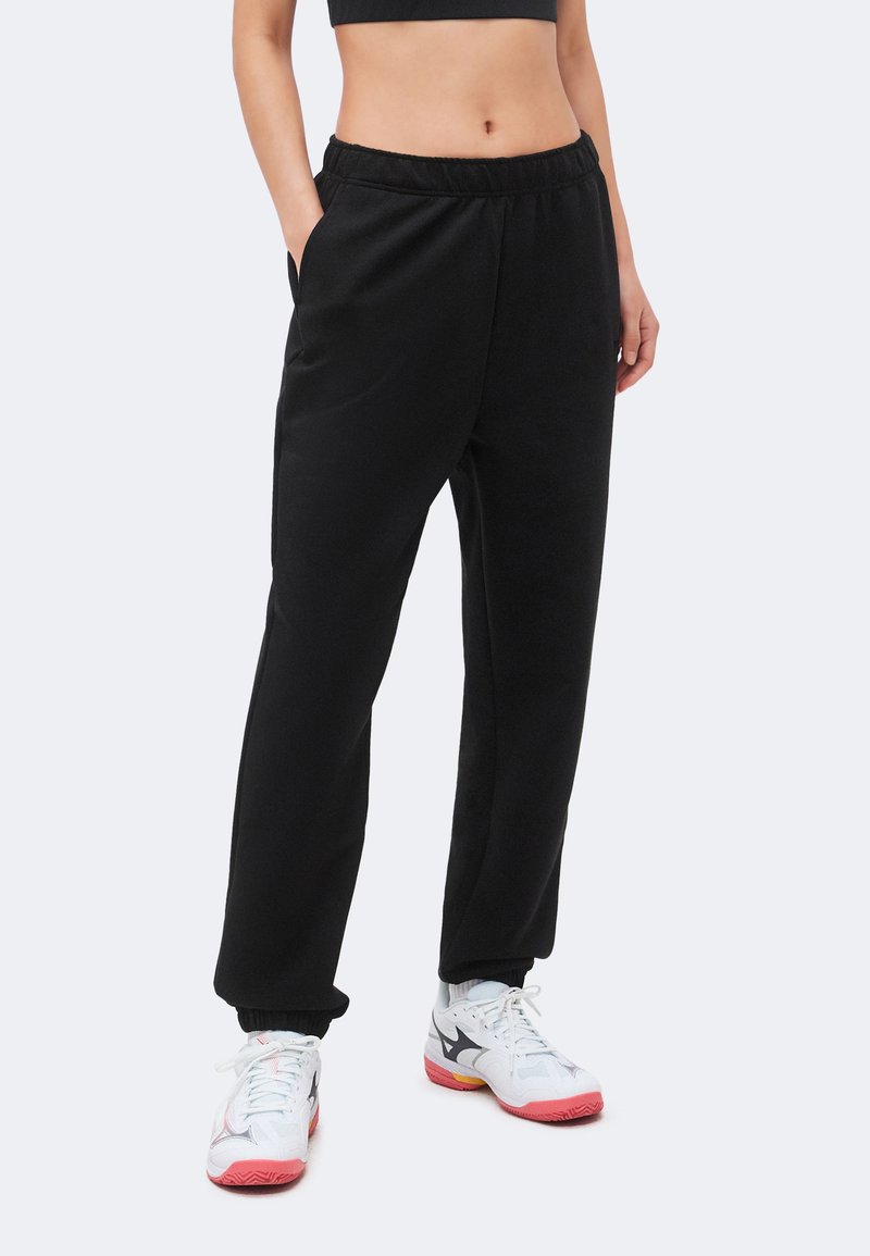 Γυναίκα που φοράει μαύρο τζόκερ με ελαστική μέση και λευκά αθλητικά παπούτσια με ροζ σόλες, όρθια με τα χέρια στις τσέπες.