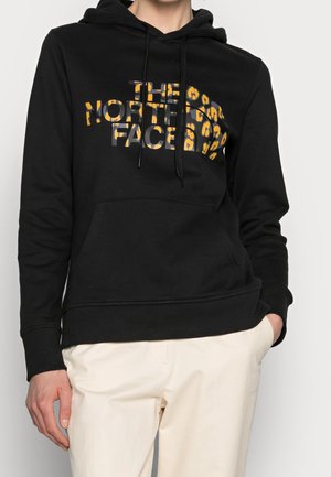 Persoon die een zwarte hoodie draagt met een gele luipaardprint "The North Face" logo, gecombineerd met lichtbeige broek, staand met één hand in de zak.