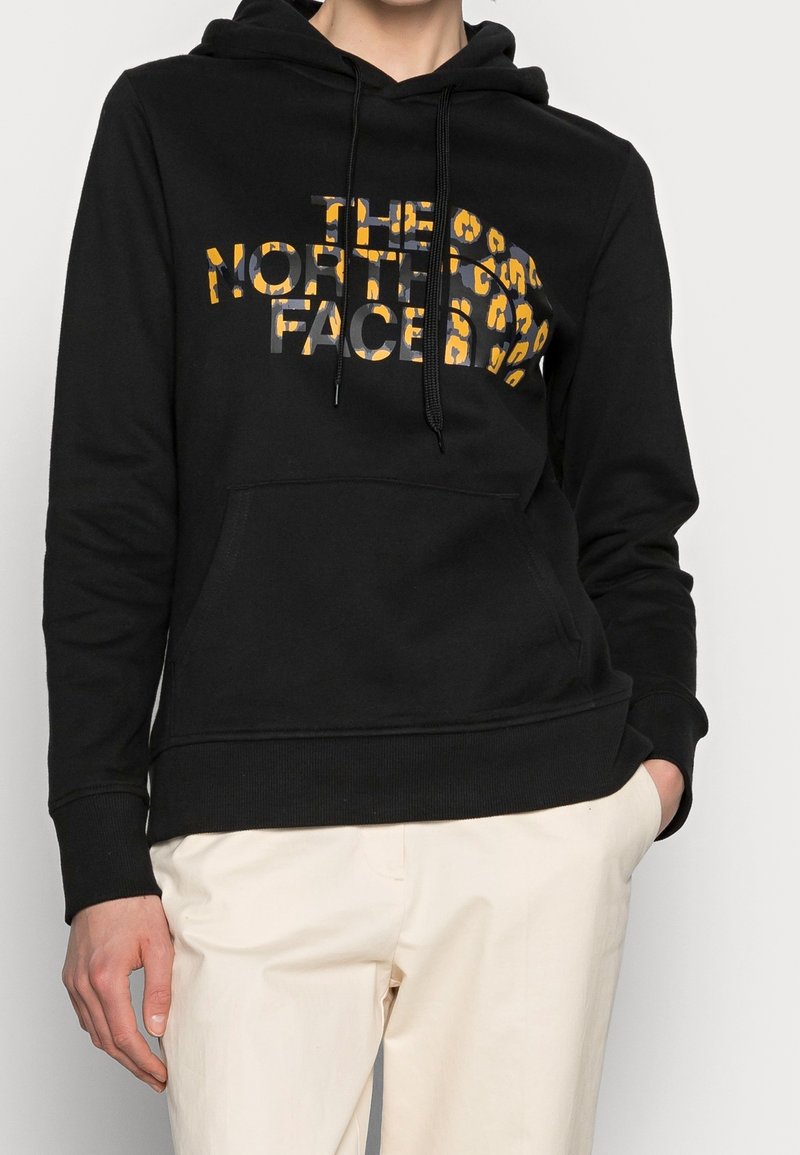 Persoon die een zwarte hoodie draagt met een gele luipaardprint "The North Face" logo, gecombineerd met lichtbeige broek, staand met één hand in de zak.