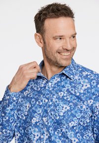 Blaues Blumenhemd mit verschiedenen Blau- und Weißtönen. Das Hemd hat einen klassischen Kragen und eine Knopfleiste, sowie eine Brusttasche.