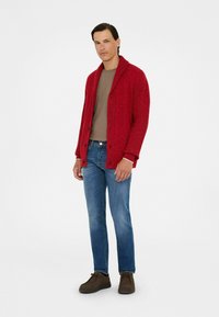Cárdigan de punto rojo con cuello de chal, usado sobre una camisa marrón, combinado con vaqueros azules y zapatos marrones. Presenta cierres de botón.