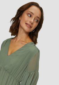 Robe verte à col en V avec de longues manches transparentes, taille froncée et texture délicate. Modèle posant avec un sourire subtil et un maquillage léger.