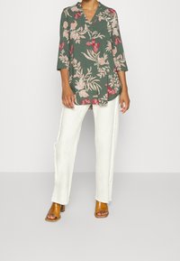 Grüne Blumenbluse mit V-Ausschnitt und Dreiviertelärmeln, verziert mit rosa und roten Blumen. Kombiniert mit weißen weiten Hosen und braunen Absatzsandalen.