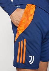 Pantaloni sportivi blu con dettagli arancioni e un design testurizzato. Caratterizzati da una tasca laterale, tre strisce verticali e un logo di branding.