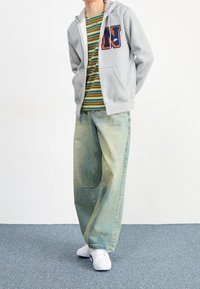 Felpa grigia con zip e patch "N" blu e arancione, t-shirt a righe verde e gialla, jeans larghi azzurri e sneakers bianche.
