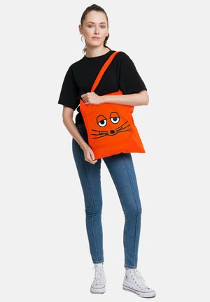 Woman wearing black shirt, blue jeans, white sneakers, holding bright orange tote bag with a relaxed cat face illustration.