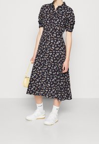 Abito midi floreale blu navy con bottoni frontali, maniche arrotolate e motivo floreale bianco, abbinato a sneakers bianche e una piccola borsa gialla.