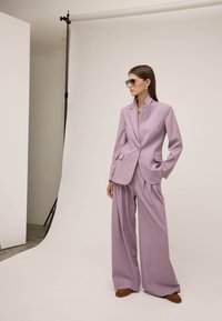 Femme portant un costume lavande oversize avec un pantalon à jambes larges et des lunettes de soleil, debout dans un studio minimaliste avec un fond blanc.