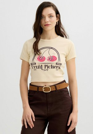 Joven con el pelo largo y castaño que lleva un top corto beige con cerezas sonrientes y el texto "Fruit Pickers", combinada con pantalones oscuros y cinturón color cuero.