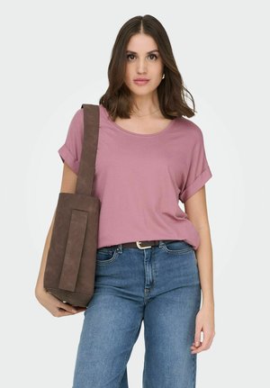 Mujer joven con cabello castaño hasta los hombros, vistiendo una camiseta malva, jeans azules y llevando un gran bolso tote marrón en el hombro.