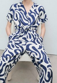 Krátký overal z bílé látky s výrazným modrým vlnkovým vzorem, stažený v pase, s límečkem a kapsami.