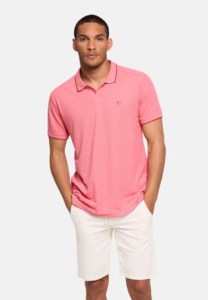 Hombre que lleva una camiseta polo rosa de manga corta y pantalones cortos blancos hasta la rodilla con las manos en los bolsillos sobre un fondo liso.