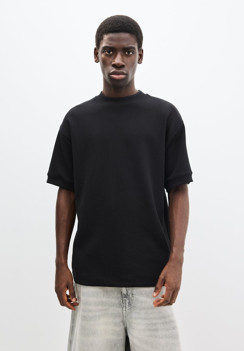 Tricou negru supradimensionat cu mâneci scurte și decolteu rotund, realizat dintr-un material neted, solid, asortat cu pantaloni scurți din denim gri deschis.