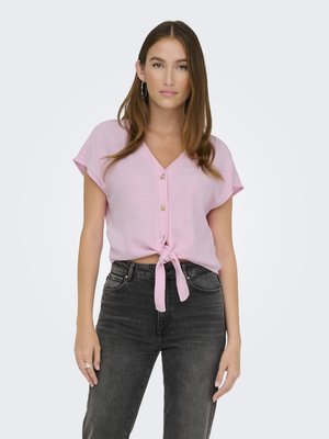 Blusa rosa a maniche corte con design a nodo frontale e chiusura con bottoni. Abbinata a jeans in denim neri. Texture liscia e stile casual.