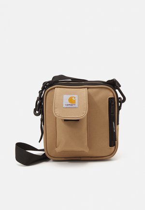 Petit sac bandoulière beige Carhartt avec poche avant à rabat, poche zippée verticale et bandoulière noire réglable sur fond uni.