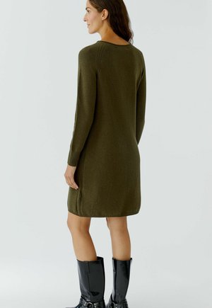 Olivgrünes Strickkleid mit langen Ärmeln, seitlichen Schlitzen und abgerundetem Saum. Das Model trägt schwarze, glänzende knielange Stiefel. Minimalistisches Design, glatte Textur.