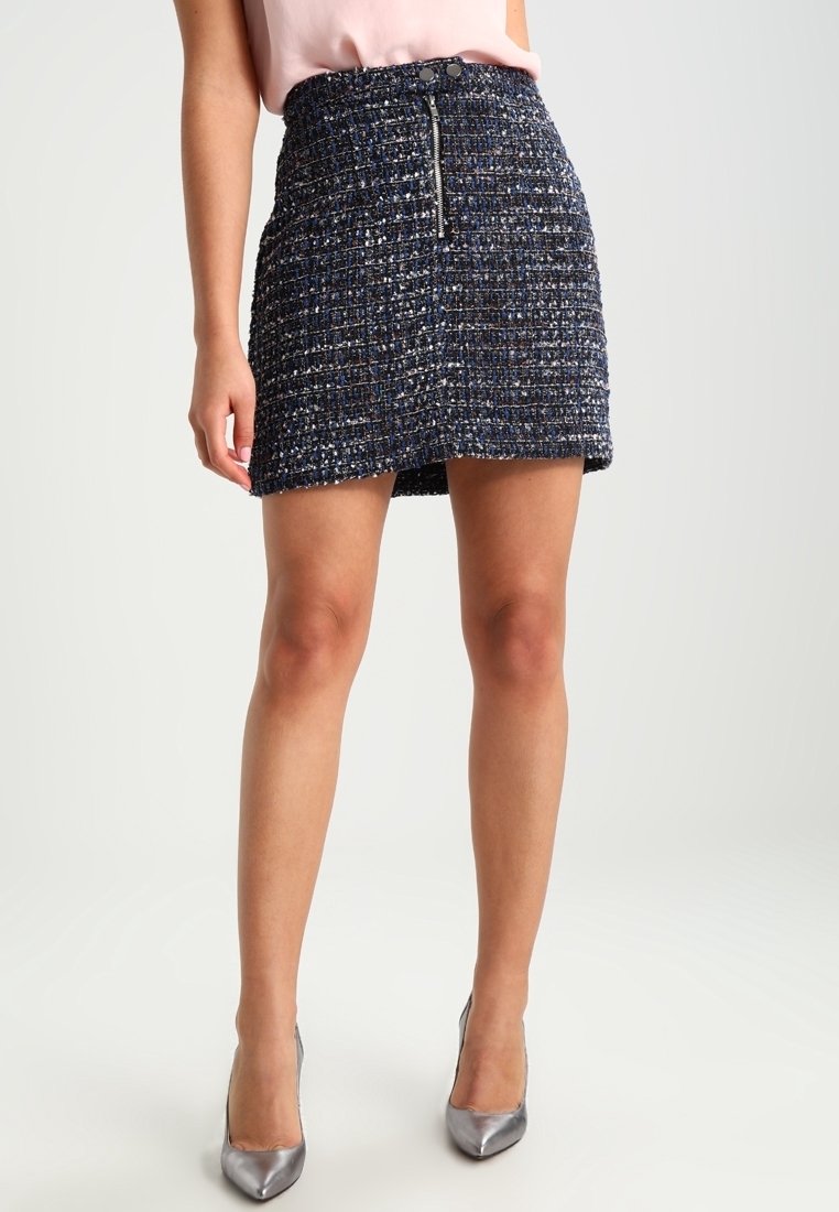 Femme portant une mini-jupe en tweed bleu marine taille haute avec fermeture éclair devant et boutons argentés, assortie à des talons pointus argentés.