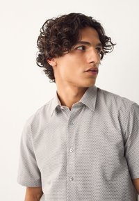 Jeune homme aux cheveux bouclés foncés portant une chemise à manches courtes gris clair à motifs, regardant sur le côté contre un fond uni.