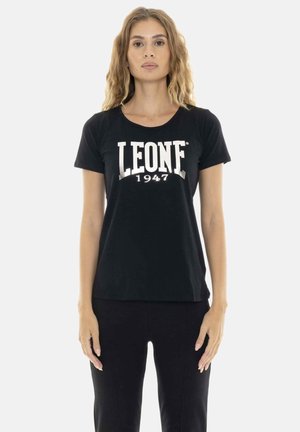 Femme aux longs cheveux bouclés portant un t-shirt noir avec le texte blanc « LEONE 1947 », debout devant un fond clair uni.