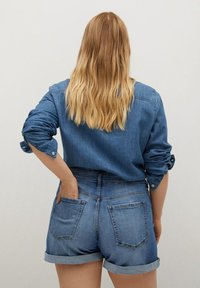 Camisa de mezclilla en azul oscuro con mangas remangadas, combinada con pantalones cortos de mezclilla azul claro que presentan una cintura alta y dobladillos doblados, destacados por los bolsillos traseros.